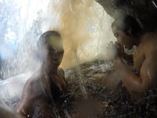 Two people enjoying a waterfall, with water splashing around them and a rocky cave in the background.