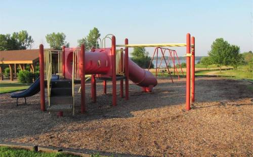 A playground featuring a red slide, swings, and a wooden structure, surrounded by grass and trees under a clear sky.