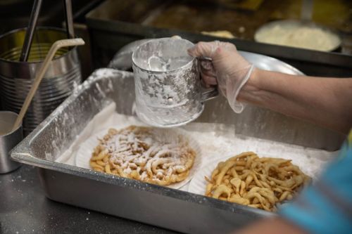A hand in a glove sprinkles powdered sugar over freshly made funnel cakes in a metal tray.