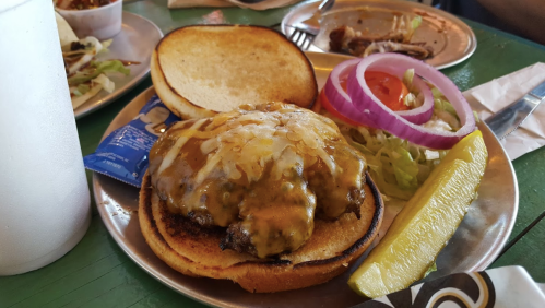 A cheeseburger with melted cheese, lettuce, tomato, and onion on a toasted bun, served with a pickle and a drink.