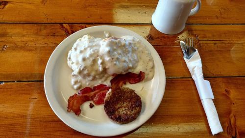 A plate of biscuits and gravy topped with bacon and a sausage patty, alongside a coffee cup and utensils.
