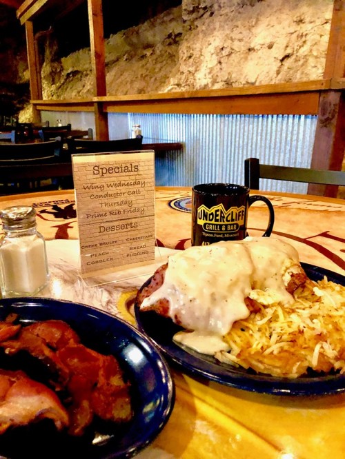 A plate of food with meat and gravy, a side of bacon, and a menu on a table in a rustic restaurant setting.