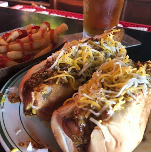 Two loaded hot dogs topped with chili and shredded cheese, served with a side of fries and a beer.