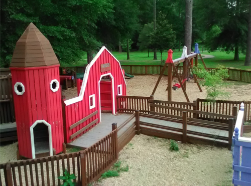 Colorful playground featuring a red barn structure, swings, and a fenced area surrounded by trees.