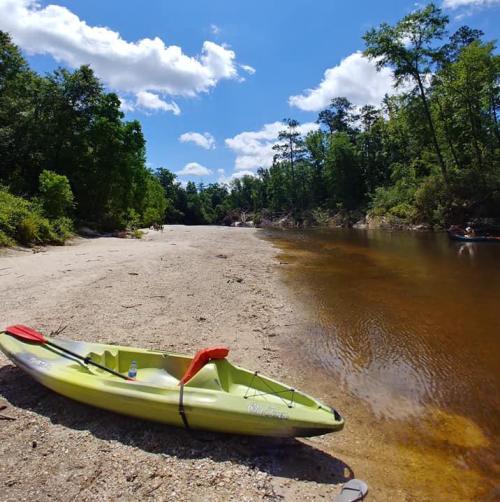 A yellow kayak rests on a sandy shore beside a calm river, surrounded by lush green trees and a blue sky with clouds.