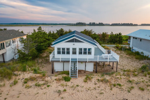 A beach house on stilts surrounded by sand and trees, with a view of the water and a cloudy sky in the background.