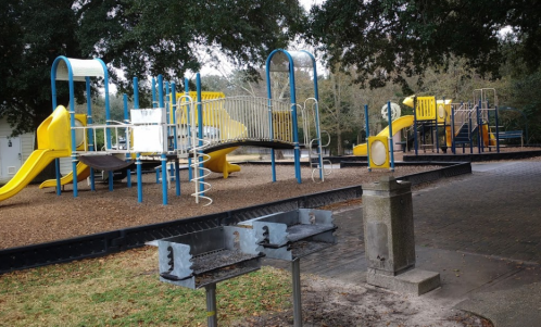 Playground with yellow and blue equipment, picnic area with grills, surrounded by trees and grass.