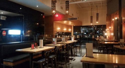 Interior of a modern restaurant with wooden tables, black chairs, and warm lighting, featuring a cozy atmosphere.