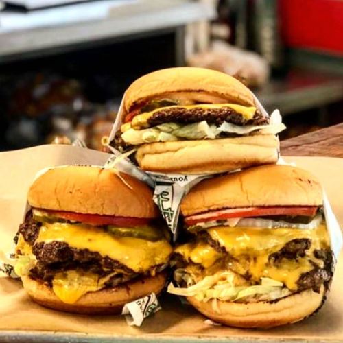 Three stacked burgers with melted cheese, lettuce, tomato, and pickles on a paper tray.