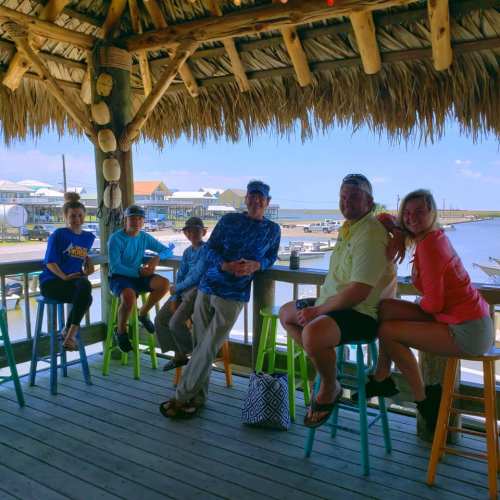 A group of six people sitting at a bar under a thatched roof, with a view of boats and water in the background.