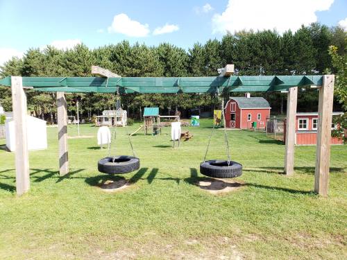 Two tire swings hang from a wooden frame in a grassy playground, with a red barn and trees in the background.