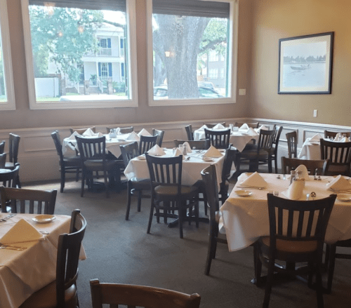 A dining room with neatly set tables, white tablecloths, and large windows overlooking a street.