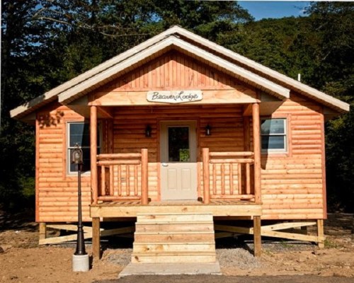 A cozy log cabin with a wooden porch, labeled "Beaver Lodge," surrounded by trees and a gravel path.