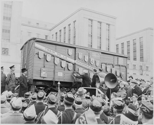 A crowd gathers around a train car during a ceremony, with banners and flags displayed in front of a large building.