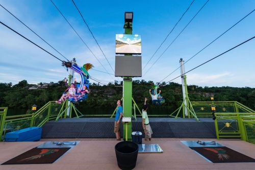 Two zip lines with riders in colorful harnesses, set against a scenic backdrop, with a monitor displaying a sky.