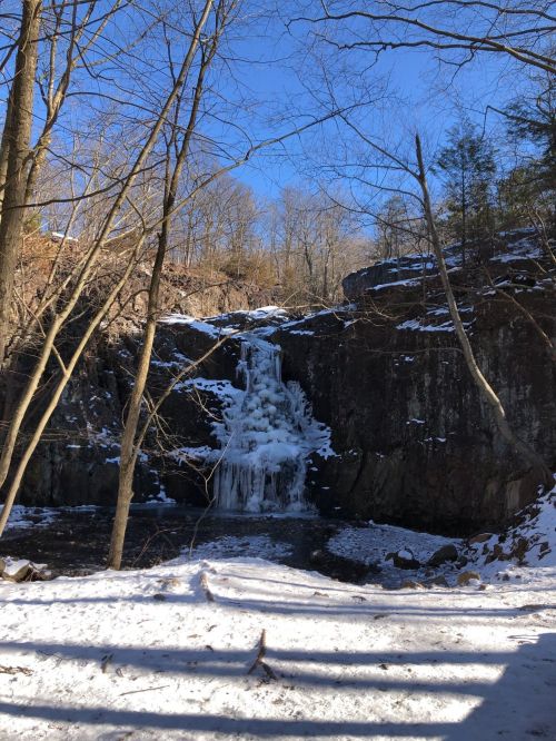 A frozen waterfall surrounded by snow-covered ground and bare trees under a clear blue sky.