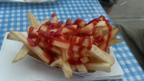 A serving of French fries drizzled with ketchup on a blue checkered tablecloth.