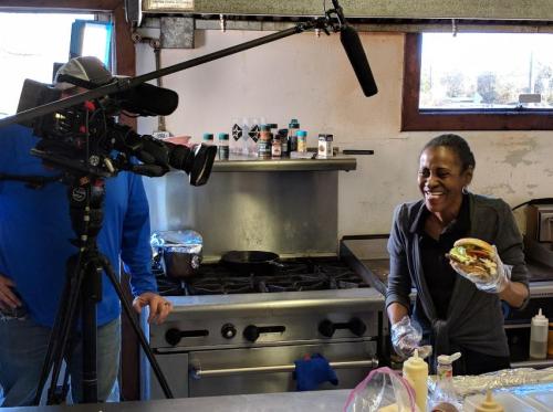 A woman in gloves holds a sandwich while smiling at a camera in a kitchen setting.