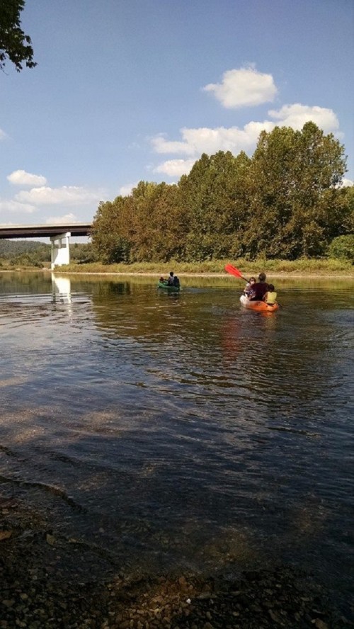 Two people kayaking on a calm river, with trees and a bridge in the background under a clear blue sky.
