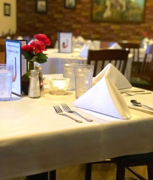 A cozy restaurant table set with white tablecloth, folded napkins, glasses, and a vase of roses.