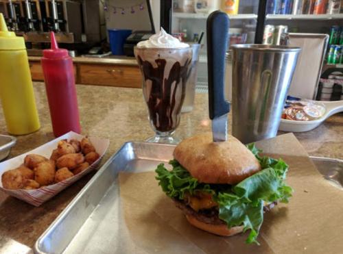 A burger with lettuce and cheese on a tray, alongside a milkshake and a basket of fried snacks with condiments.