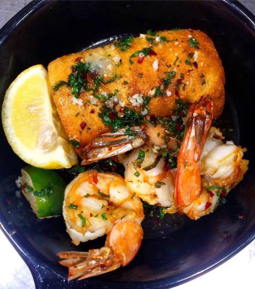 A skillet with a golden-brown breaded item, shrimp, and lemon wedges, garnished with herbs and spices.