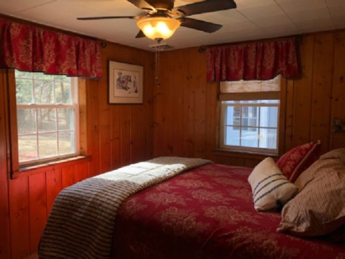 Cozy bedroom with wooden walls, a red bedspread, and sunlight streaming through two windows.