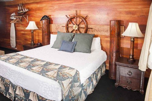 Cozy bedroom with a wooden wall, a ship's wheel decor, a queen bed, and bedside lamps. Nautical-themed bedding.