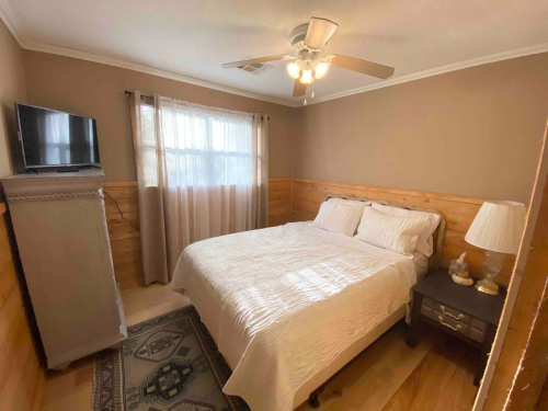 Cozy bedroom with a white bed, wooden accents, a TV, and a lamp, featuring natural light from a window.