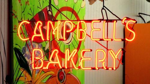 Neon sign reading "Campbell's Bakery" against a colorful, artistic background.