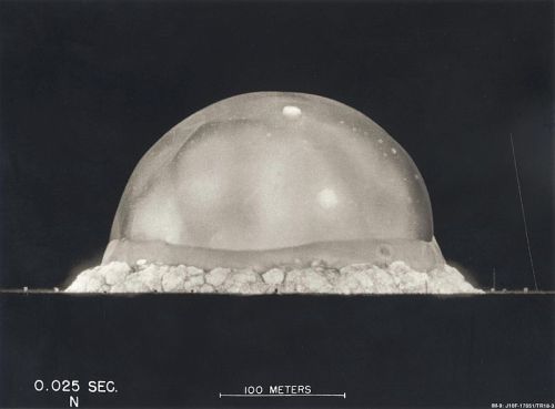 A black and white image of a nuclear explosion, showing a large mushroom cloud rising above the ground.