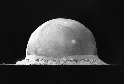 A black and white image of a large, dome-shaped cloud, resembling an explosion, rising from the ground.