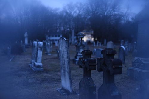 A foggy graveyard at dusk, featuring old gravestones and crosses surrounded by trees.