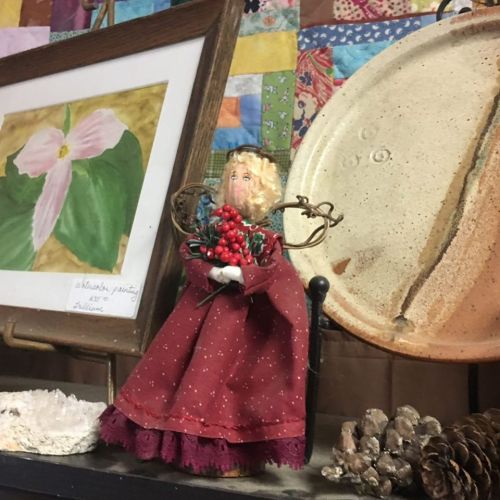 A decorative doll in a red dress holds berries, surrounded by art and rustic items on a shelf.