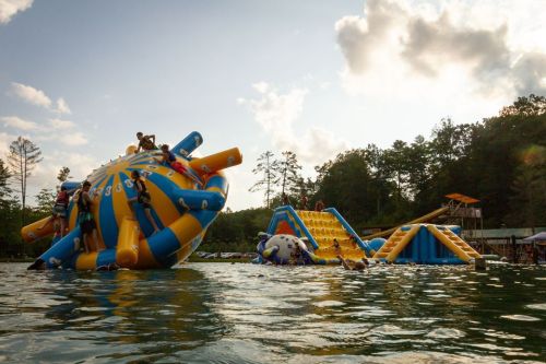 A colorful inflatable water playground with people climbing and sliding, set against a sunny sky and trees.