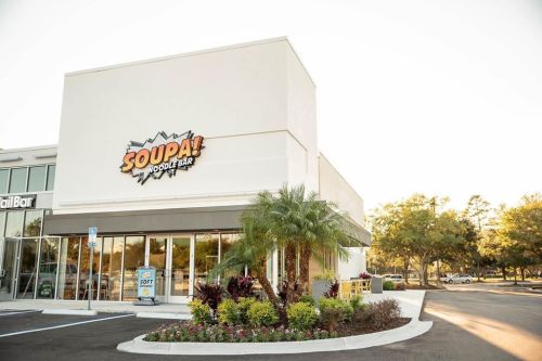 Exterior of a modern noodle bar named "SOUPA!" with colorful landscaping and a clear sky in the background.
