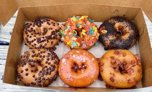 A box of six assorted donuts: chocolate chip, fruity cereal, maple bacon, and more, on a wooden surface.