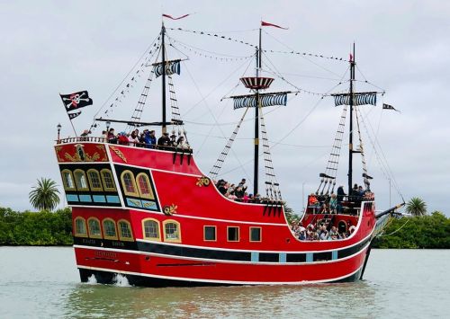 A colorful pirate ship with sails and flags, sailing on calm waters with people on board and greenery in the background.