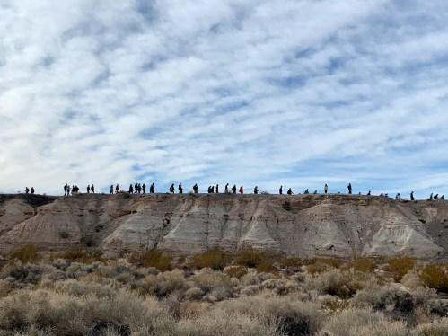 A line of people walking along a ridge against a cloudy sky, with desert vegetation in the foreground.