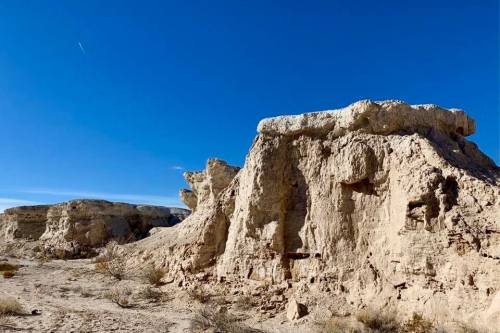 A rugged desert landscape featuring large rock formations under a clear blue sky. Sparse vegetation is visible.