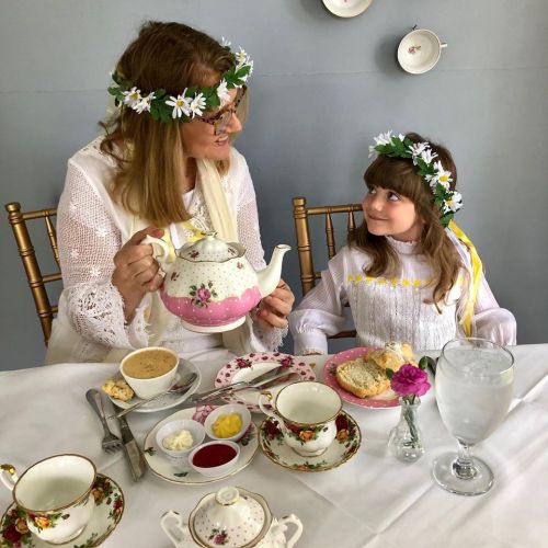 A woman and a girl share tea at a beautifully set table, both wearing floral crowns and smiling at each other.