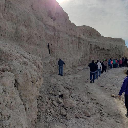 A group of people walking along a rocky terrain with steep, eroded cliffs in the background under a cloudy sky.