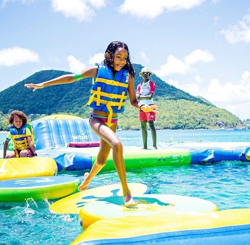 A girl in a life jacket jumps on a floating platform in a sunny water park, with mountains in the background.