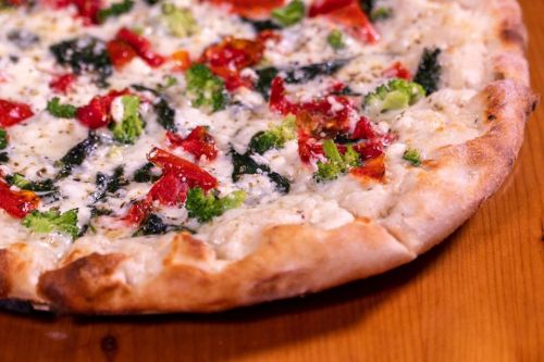 Close-up of a pizza topped with cheese, red peppers, and broccoli on a wooden table.