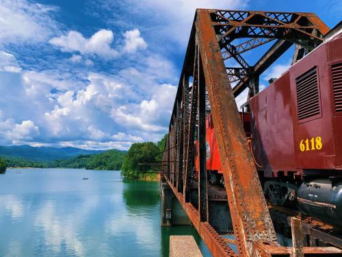 A rusty train bridge spans a calm lake, with mountains and fluffy clouds in the background.
