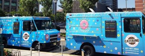 Two colorful food trucks with blue exteriors and donut designs, parked in an urban area, promoting catering services.