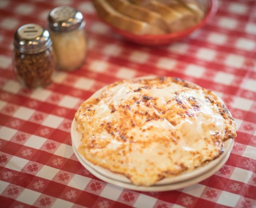 A cheesy pizza on a white plate, set on a red and white checkered tablecloth, with condiments in the background.