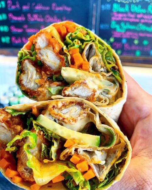 A hand holding a cut wrap filled with chicken, lettuce, carrots, and sauce, with a colorful menu in the background.
