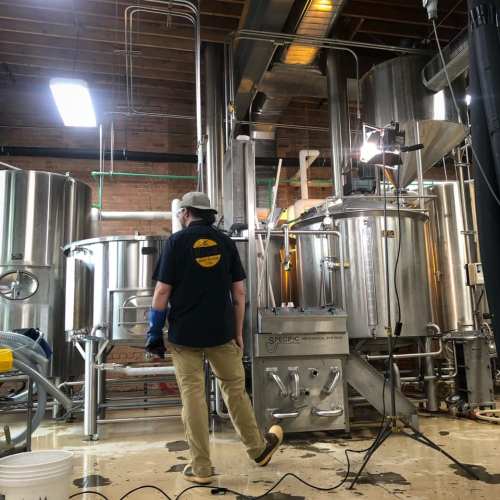 A person in a black shirt and cap stands in a brewery, surrounded by large stainless steel brewing equipment.