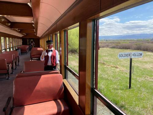 A train conductor stands inside a vintage train car, with a view of fields and a "SOLDIER HOLLOW" sign outside.